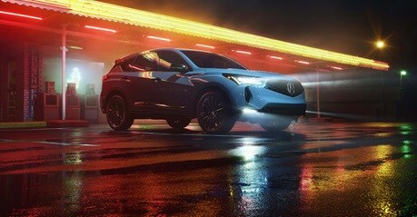 Acura RDX 2026 en Urban Gray Pearl estacionada frente a un restaurante iluminado con neón de noche (SUV crossover deportiva prémium).